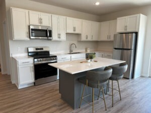 Modern kitchen with white cabinets, stainless steel appliances, a center island with a gray base, two gray and gold stools, wood-look flooring, and minimal decor. Bright natural light fills the space.