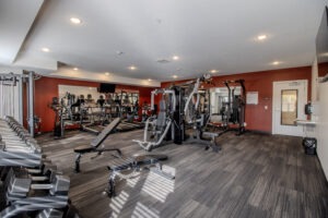 A modern gym with various exercise machines, free weights, benches, and cardio equipment, set against a red accent wall with mirrors and bright overhead lighting.
