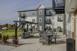 A modern apartment building with gray and white siding, featuring an outdoor patio area with dining tables, chairs, grills, a pergola, and landscaped surroundings on a sunny day.