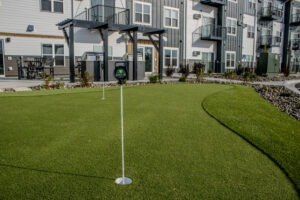 A putting green with two golf holes and flagsticks is set in front of a modern apartment building with balconies and stairs. Landscaping with rocks and plants surrounds the area.