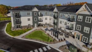 Aerial view of a modern apartment building with black and white exterior, balconies, a small putting green, seating area, pergola, and surrounding landscaping on a sunny day.