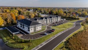 Aerial view of a large, modern apartment building surrounded by trees with autumn foliage, parking lots with several cars, and a winding road under a clear sky.