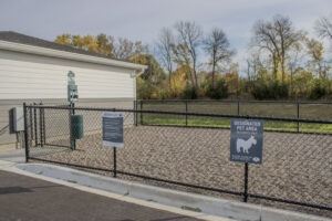A fenced designated pet area with gravel ground, signs, and a waste station, located next to a building. Trees with autumn foliage are visible in the background.