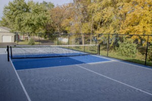 Outdoor pickleball court with a blue playing surface and white lines, surrounded by a black chain-link fence. Trees with green and yellow autumn leaves are visible in the background.