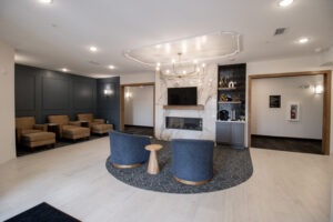 A modern lobby with blue chairs, brown armchairs, a round wooden table, a fireplace, a TV above it, decorative shelves, and stylish lighting. The floor is light tile with a dark circular accent area.