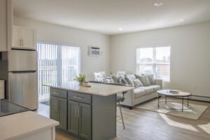 Bright, modern open-concept living space with a kitchen island, stainless steel fridge, white cabinets, dining table, light gray sofa, round coffee table, large windows, and sliding glass doors letting in natural sunlight.