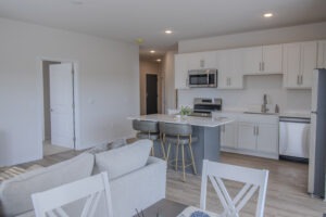 Modern kitchen and living area with white cabinets, stainless steel appliances, an island with two barstools, a light gray sofa, and a dining table with white chairs in a bright, open-plan apartment.