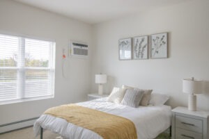 A bright bedroom with a large window, a bed with white sheets and a yellow throw blanket, two bedside tables with lamps, and three botanical prints hanging above the bed. An air conditioner is mounted on the wall.