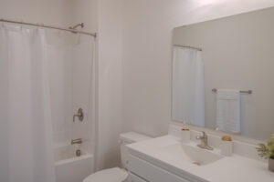 A clean, modern bathroom with a white shower curtain, bathtub, toilet, large mirror, and white vanity with a sink. Minimalist decor includes soap dispensers and a small plant on the counter.