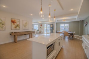 Bright open-concept kitchen and living area with white island, hanging pendant lights, modern art on the walls, shuffleboard table, dining table, and large windows letting in natural light.