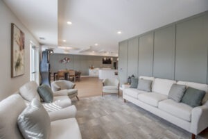 Modern living room with light gray sofas and armchairs, green accent pillows, a patterned rug, and a view of an open dining area and kitchen in the background. Walls are light green panels with a large window on one side.
